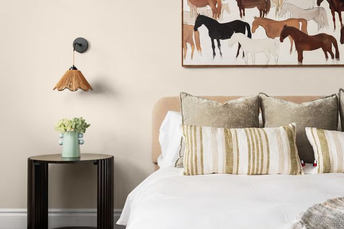Cozy modern bedroom interior with beige upholstered headboard, white bedding, striped lumbar pillows and velvet cushions, framed horse artwork above the bed, rattan wall sconce, and a round black bedside table holding a mint vase with flowers.