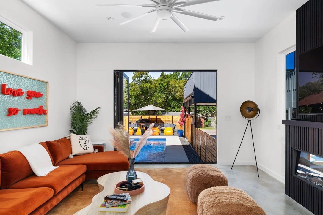 Bright modern living room with a burnt-orange sectional and white walls, folding glass doors opening to a sunny backyard pool and patio with yellow lounge chairs and umbrella.
