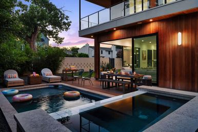 Inviting modern backyard patio at dusk with a rectangular pool holding colorful inflatable rings, wood-deck dining table and lounge chairs, warm wood-paneled house with large sliding glass doors, surrounding greenery and a purple-pink sunset sky.