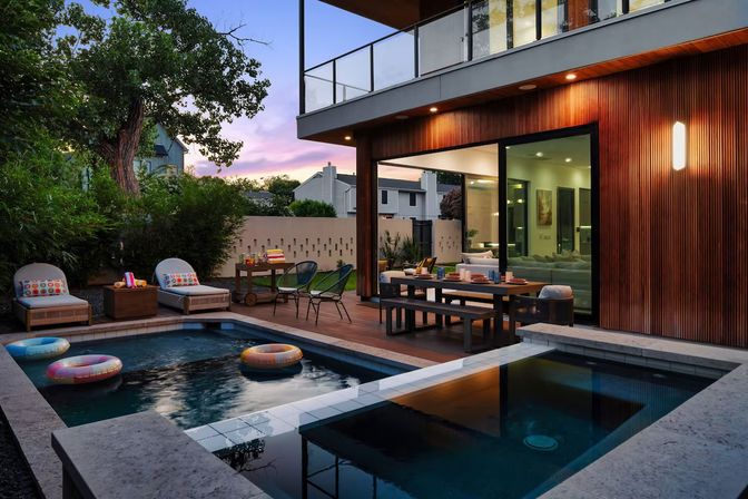 Inviting modern backyard patio at dusk with a rectangular pool holding colorful inflatable rings, wood-deck dining table and lounge chairs, warm wood-paneled house with large sliding glass doors, surrounding greenery and a purple-pink sunset sky.