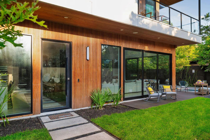 Sunlit modern wood-clad home with floor-to-ceiling sliding glass doors opening to a backyard patio, green lawn, outdoor lounge chairs with yellow cushions, and an upper balcony.