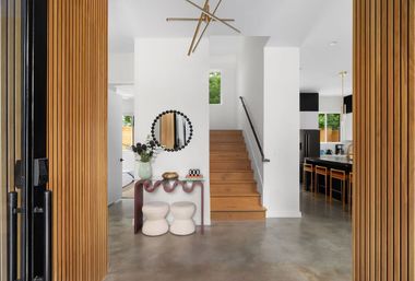 Modern entryway with wooden slat pivot doors opening to a polished concrete foyer, wavy console with vase and round mirror, wooden staircase, and open-plan kitchen with bar stools.