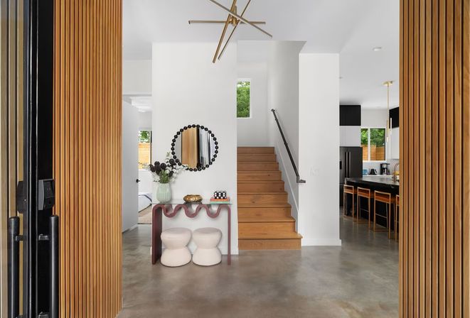 Modern entryway with wooden slat pivot doors opening to a polished concrete foyer, wavy console with vase and round mirror, wooden staircase, and open-plan kitchen with bar stools.