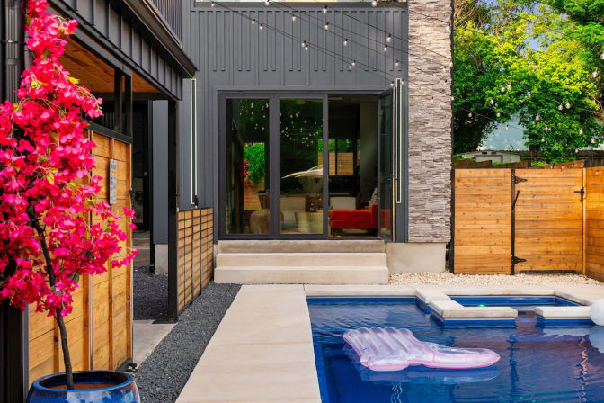 Modern backyard patio with a rectangular pool and bright pink bougainvillea, a clear inflatable float on blue water, concrete steps to glass sliding doors, wooden privacy fence and string lights.
