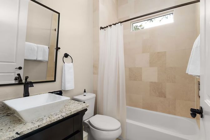 Bright modern bathroom with granite countertop, rectangular vessel sink, black fixtures, toilet and bathtub with beige tile and white shower curtain.