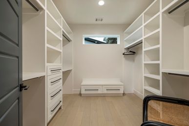 Modern minimalist walk-in closet in a contemporary home with white built-in shelving and drawers, light hardwood floors, a small high window, black door trim and a woven chair