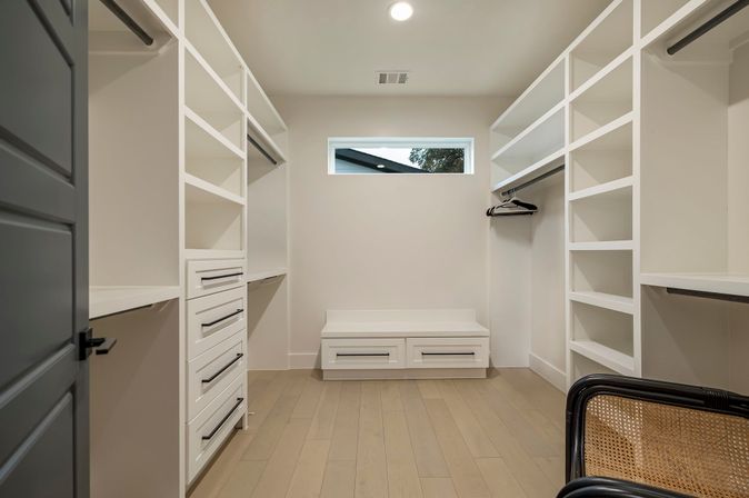 Modern minimalist walk-in closet in a contemporary home with white built-in shelving and drawers, light hardwood floors, a small high window, black door trim and a woven chair