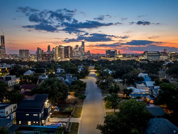 Aerial sunset view down a tree-lined residential street leading to the Austin, Texas skyline with glowing downtown skyscrapers under an orange and blue sky.