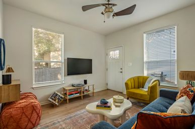 Bright modern living room in a suburban home with a wall-mounted TV, mustard yellow accent chair, blue sofa with colorful pillows, round white coffee table, woven pouf, hardwood floors, ceiling fan, and two large windows with blinds.