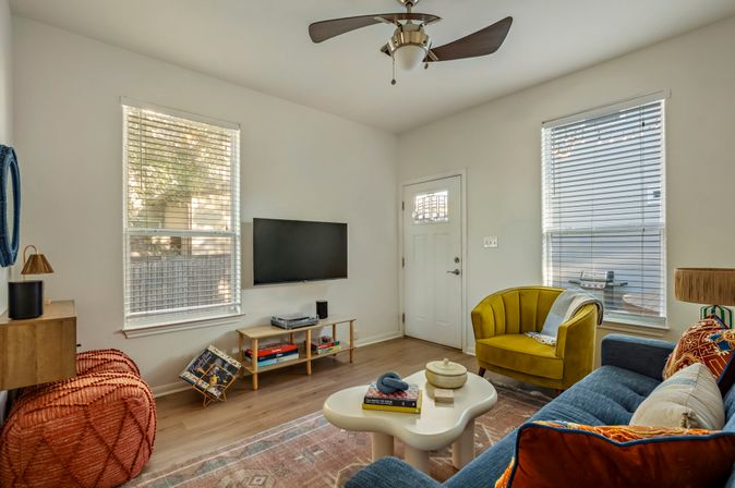 Bright modern living room in a suburban home with a wall-mounted TV, mustard yellow accent chair, blue sofa with colorful pillows, round white coffee table, woven pouf, hardwood floors, ceiling fan, and two large windows with blinds.