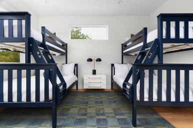 Bright bunk bedroom with two navy-blue double bunk beds (four beds total), crisp white linens, white nightstand with a black dual-lamp, small horizontal window, and blue-green checkered rug.