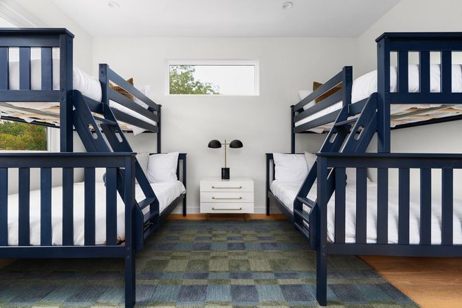 Bright bunk bedroom with two navy-blue double bunk beds (four beds total), crisp white linens, white nightstand with a black dual-lamp, small horizontal window, and blue-green checkered rug.