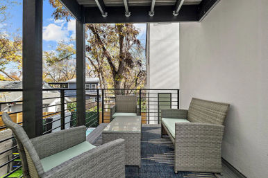 Modern covered apartment balcony with gray wicker seating and glass coffee table overlooking a tree-lined residential street.