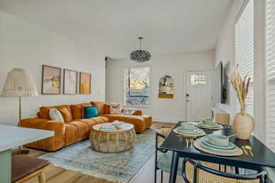Sunlit modern open-plan living and dining room with caramel tufted sectional, boho rattan coffee table, patterned rug, teal accent pillows and a set dining table.