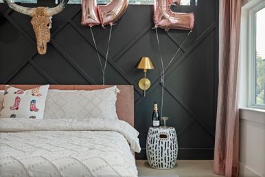 Boho-chic modern bedroom with a black geometric accent wall, blush pink upholstered bed, white textured bedding and cowboy-boot print pillow, rose-gold balloons, decorative longhorn skull, gold wall sconce, patterned ceramic bedside table with a champagne bottle and coupe glass, and pink velvet curtains by a window.