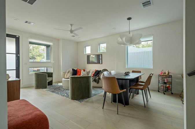 Bright modern open-concept living and dining room with light wood floors, green velvet lounge chairs, a sofa with colorful throw pillows on a zebra-print rug, black dining table with tan leather chairs, sculptural white pendant light and large windows.