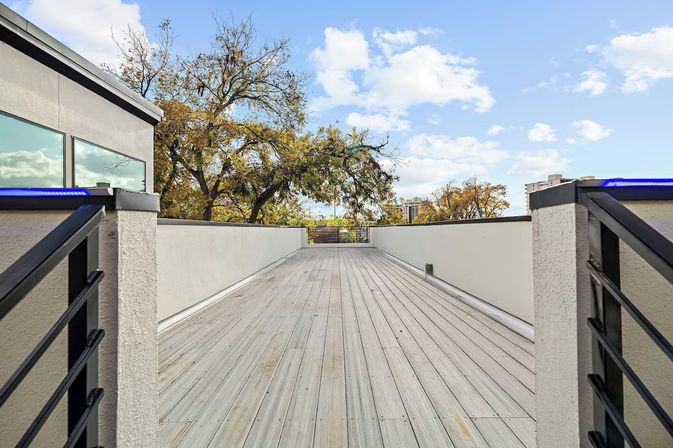 Modern urban rooftop deck with light wood plank flooring, low white parapet walls and black metal railings with blue LED accents, overlooking leafy trees and a distant city skyline under a bright blue sky