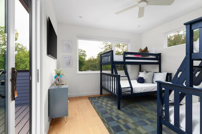 Bright modern bedroom with navy twin-over-full bunk beds, hardwood floors, sliding glass door to balcony, large windows showing tree-lined outdoor views and patterned area rug