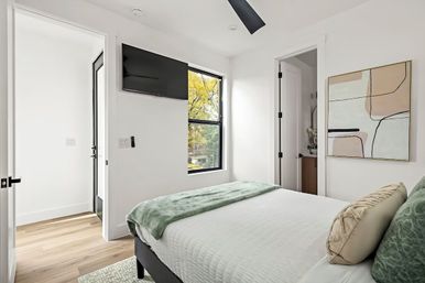 Bright modern minimalist bedroom with white walls, wall-mounted TV, large window showing tree-lined view, bed with green throw blanket and abstract wall art.