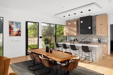 Sunlit modern open-plan kitchen and dining area with a wooden table and leather chairs, marble island with bar stools, pendant lights, and large glass doors to a green backyard.