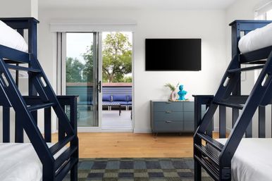Bright modern bedroom with navy bunk beds, hardwood floors, a blue dresser and wall-mounted TV, sliding glass door opening to a deck with outdoor seating and trees beyond.