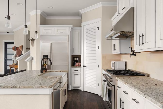 Bright modern farmhouse-style kitchen featuring a sprawling granite island, white shaker cabinets, stainless steel fridge and range, glass pendant lights, and dark hardwood floors.