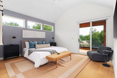 Modern bedroom with vaulted ceiling and chevron accent wall featuring three narrow windows, king bed with blue pillows, tan bench on a beige area rug, sliding glass door to a balcony overlooking leafy neighborhood