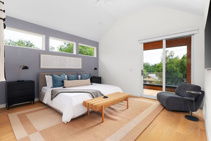Modern bedroom with vaulted ceiling and chevron accent wall featuring three narrow windows, king bed with blue pillows, tan bench on a beige area rug, sliding glass door to a balcony overlooking leafy neighborhood