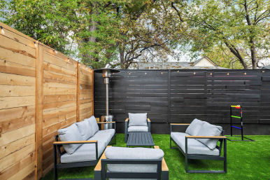 Cozy modern backyard patio with gray cushioned outdoor lounge set on green turf, black horizontal fence and natural wood privacy wall, string lights, patio heater and ladder toss game.