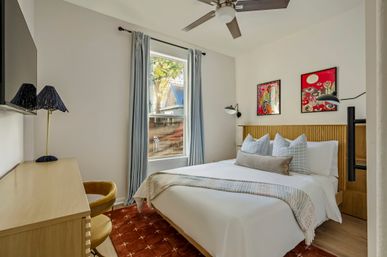 Bright, cozy bedroom interior with queen bed and ribbed light-wood headboard, white linens and striped throw, three decorative pillows, blue curtains framing a sunny window, ceiling fan, wooden desk with navy lamp and mustard swivel chair, colorful framed art on the wall.