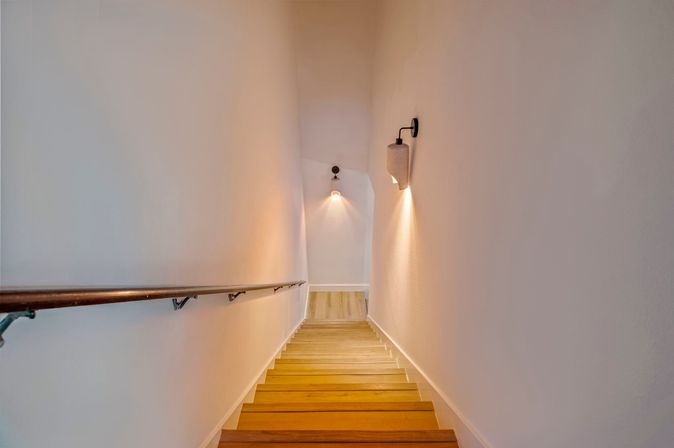 Modern narrow residential staircase with wooden steps, white walls, a metal handrail on the left and warm wall sconces casting light down to a small landing.