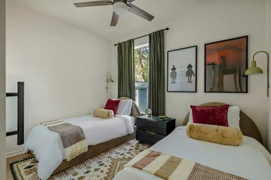 Cozy modern twin bedroom with two single beds in white linens, velvet red accent pillows, faux-fur bolsters, green curtains, black nightstand, framed wall art, ceiling fan and patterned rug.
