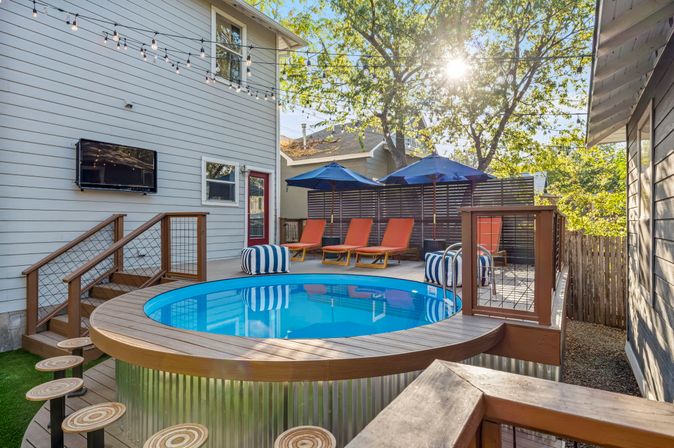 Sunny modern suburban backyard pool deck with round above-ground pool, orange lounge chairs under blue umbrellas, striped poufs, string lights and wall-mounted outdoor TV