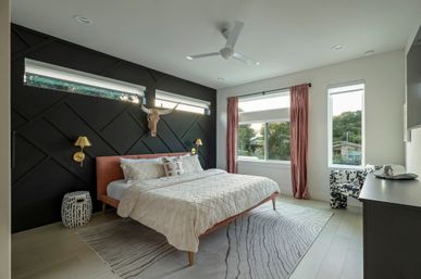 Modern master bedroom interior with black geometric accent wall, blush-upholstered bed, decorative longhorn skull, brass wall sconces, large windows with pink curtains, patterned area rug, ceiling fan and light wood floors with a suburban view