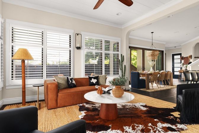 Sunlit open-plan living and dining room with tan leather sofa, round white marble coffee table, cowhide and jute rugs, white plantation shutters, rattan dining chairs, indoor cactus and natural wood accents.