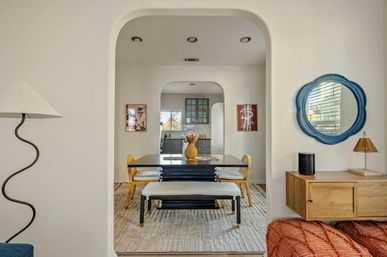 Light-filled open-concept dining room framed by an arched doorway — black table with bench and woven chairs, decorative vase centerpiece, blue round wall mirror, and view into a gray kitchen