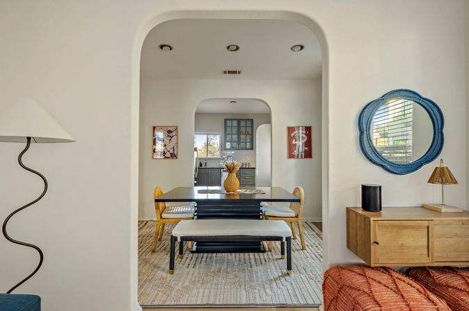 Light-filled open-concept dining room framed by an arched doorway — black table with bench and woven chairs, decorative vase centerpiece, blue round wall mirror, and view into a gray kitchen