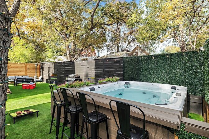 Residential backyard patio with a raised hot tub and four black bar stools, artificial turf, lounge seating, grill and cornhole game under mature shade trees and string lights