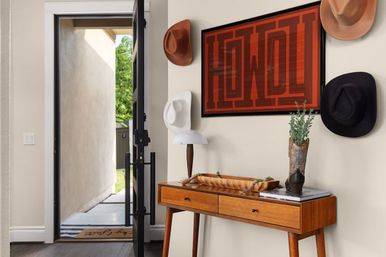 Bright western-themed entryway with open black-framed front door, mid-century wooden console table holding a lamp and tray, red “HOWDY” wall art, hanging cowboy hats, and a decorative cowboy boot vase with greenery overlooking a sunlit yard.
