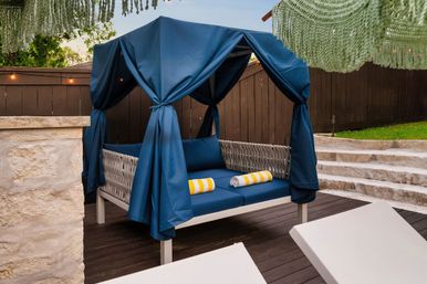 Cozy navy-canopy outdoor daybed with woven frame and blue cushions, two yellow-and-white striped rolled towels on a dark wooden deck by stone steps and a brown fence — backyard patio lounge.