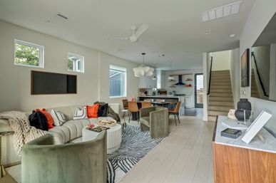 Modern open-concept living room and kitchen interior with light hardwood floors, cream sectional sofa with orange and black accent pillows, green velvet chairs, round white coffee table on a zebra rug, dining table with leather chairs, pendant lights, staircase and natural light from windows.