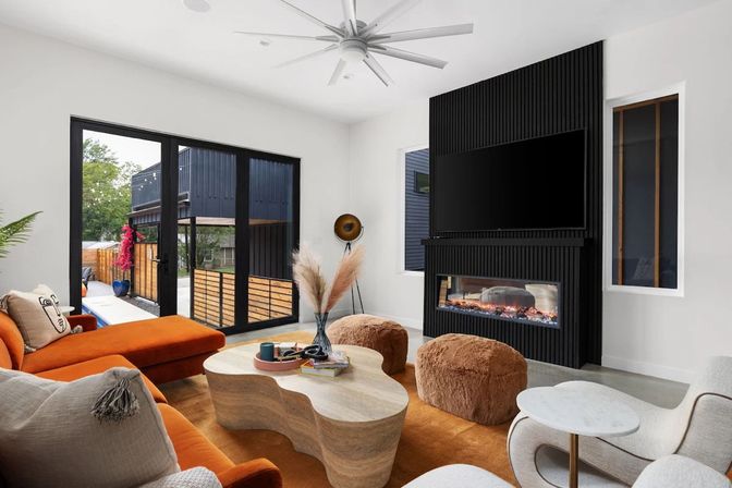 Bright modern living room with orange sectional, sculptural stone coffee table, fuzzy brown poufs, wall-mounted TV above a linear fireplace framed in black fluted paneling, large black-framed sliding doors opening to a patio.