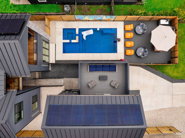 Aerial view of a modern residential courtyard with a rectangular blue pool and spa, gray wood deck with orange loungers and umbrella, rooftop solar panels, seating area and small lawn