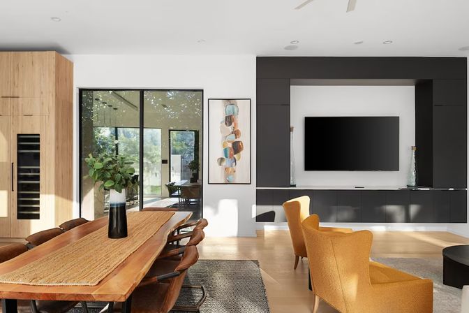 Sunlit modern open-plan living and dining room with a live-edge wooden dining table and leather chairs, mustard-yellow lounge chairs, built-in black media wall with flat-screen TV, vase of greenery on the table, and large sliding glass doors opening to a leafy backyard.