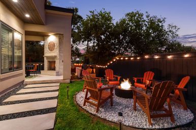 Cozy backyard patio at dusk with a circular fire pit surrounded by red Adirondack chairs on white gravel, string lights along a dark fence, stone walkway to a covered porch with an outdoor fireplace and seating, and a grassy lawn with trees.