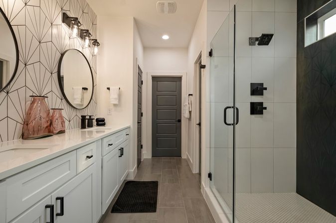 Sleek modern master bathroom with long white double vanity, round mirrors and geometric accent wall, glass-enclosed walk-in shower with matte black fixtures, gray tile floor and dark gray paneled door