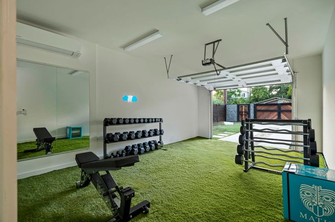 Bright residential garage gym with green turf flooring, adjustable bench, mirrored wall, two dumbbell racks and an open garage door leading to a backyard