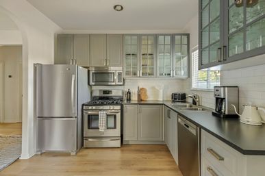 Bright modern kitchen with gray cabinets and glass-front uppers, stainless steel refrigerator, gas range and microwave, dishwasher, black countertops, white subway tile backsplash, coffee maker and kettle on light hardwood floors.