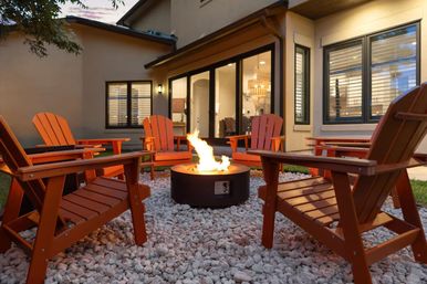 Cozy suburban backyard patio at dusk with a lit round fire pit surrounded by red Adirondack chairs on white decorative gravel, framed by sliding glass doors and warm indoor lighting.