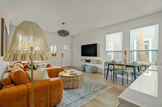 Bright modern boho open-plan living room with a burnt-orange sectional sofa, tasseled rattan floor lamp and round wicker coffee table on a blue patterned rug, wall-mounted TV, dining table with green chairs by sunlit windows and light hardwood floors.